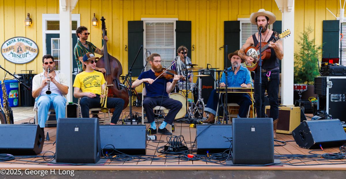 A band performs on an outdoor stage at the Abita Springs Busker Festival, with musicians playing guitar, fiddle, upright bass and drums in front of a yellow building.