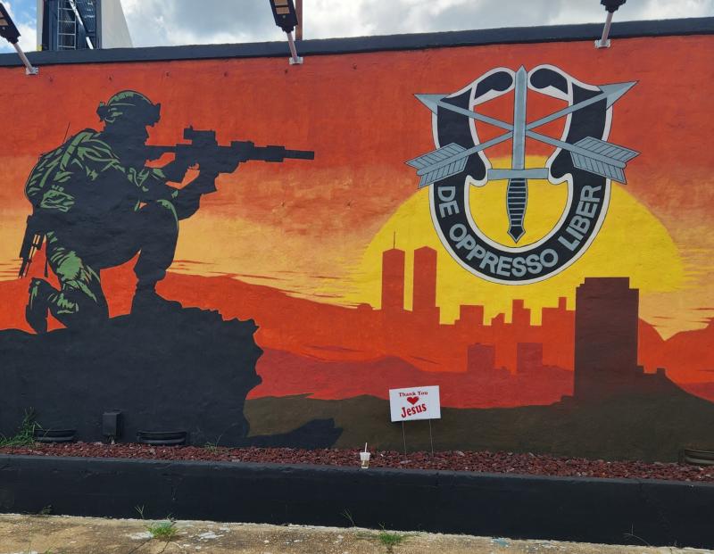 A mural painted on the side of a building shows a kneeling Special Forces soldier aiming a rifle, silhouetted against a vivid orange and yellow sunset skyline. To the right, a large “De Oppresso Liber” U.S. Army Special Forces emblem is featured prominently.