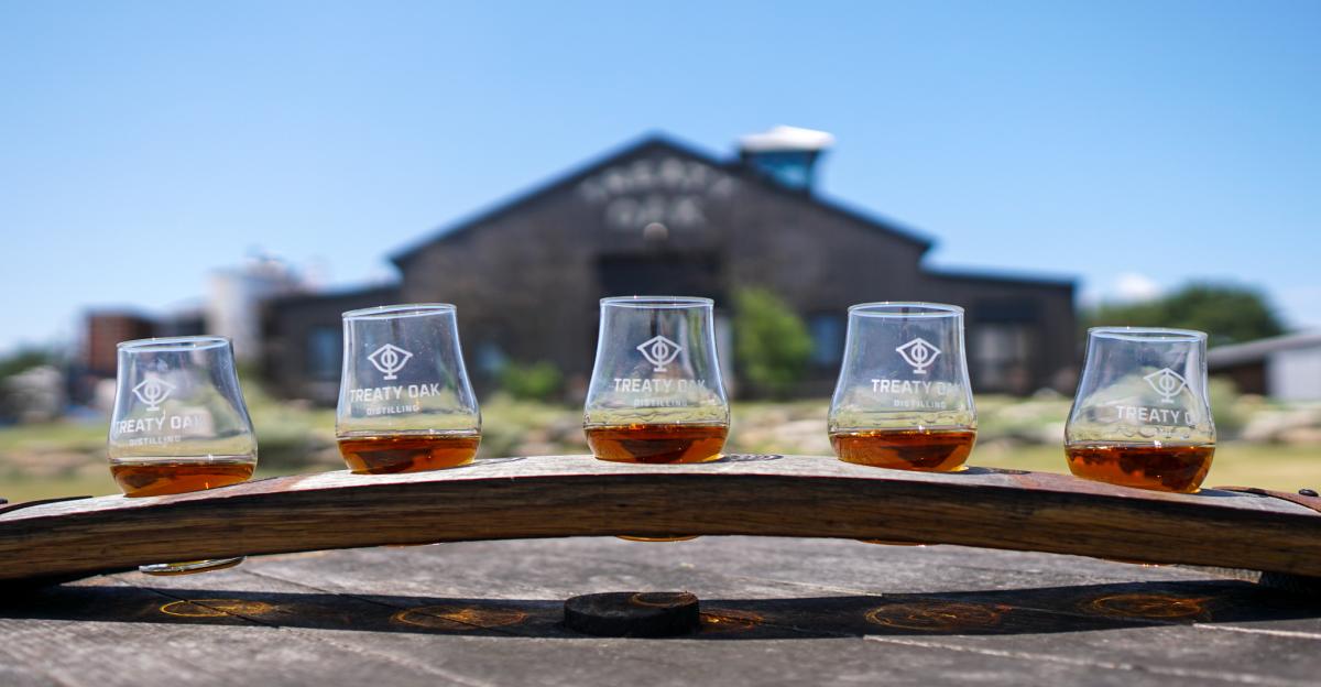Image of five short tasting glasses sitting on a wooden plan on a table with the large Treaty Oak Distilling barn in the background.