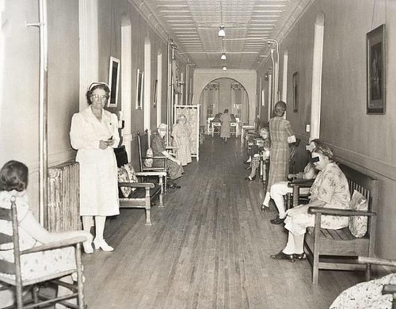 black and white photo of a hospital hallway with patients and a nurse from the early 1900s