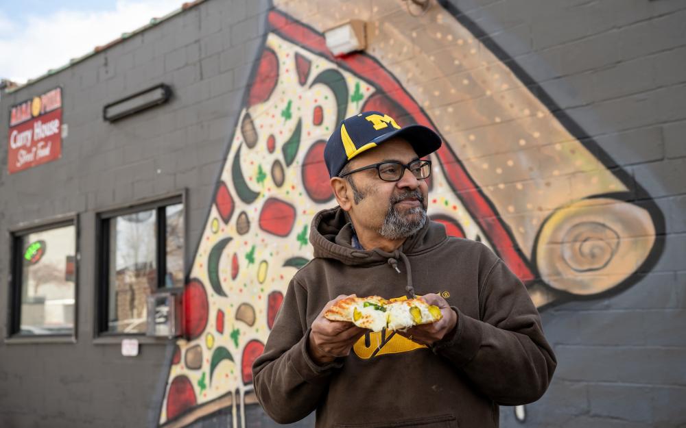 Man stands outside of building, in front of a large mural of a slice of pizza. He's holding a large slice of pizza.