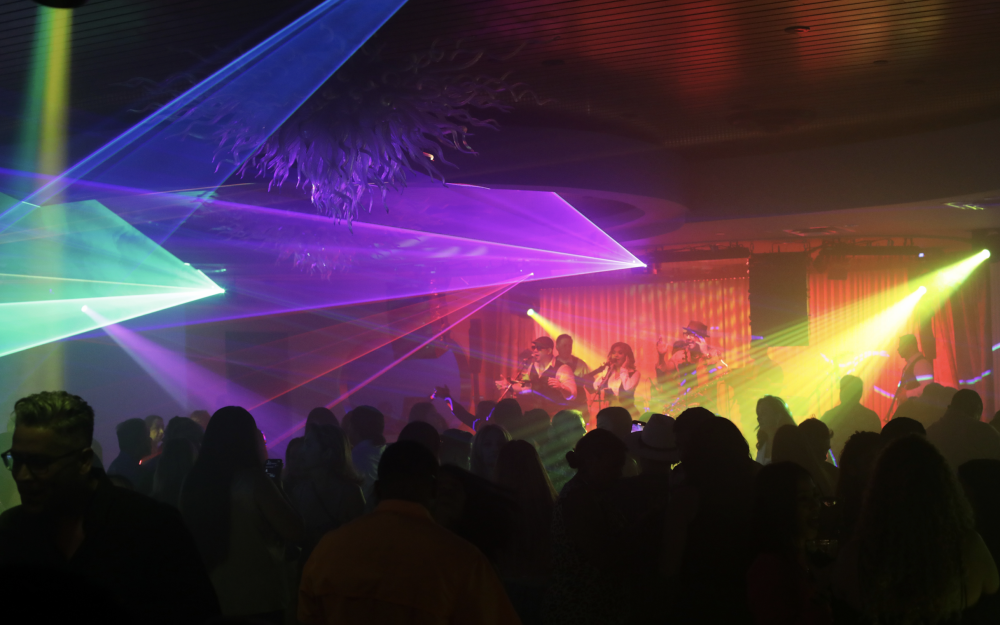 Shadows of people on a crowded dance floor under colorful gobo lights while a band plays on stage.