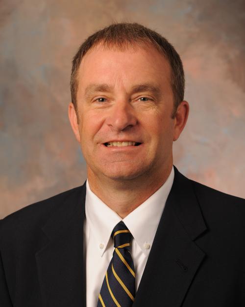 Professional headshot of a man in a dark suit, white shirt, and navy striped tie, smiling in front of a soft, multicolored studio backdrop