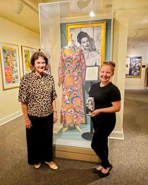 Ava Thompson and Dominique Benedict pose in front of a 1960s era dress of Ava Gardner's with a psychdelic pattern. Dominique holds a bag of the Ava Gardner coffee.