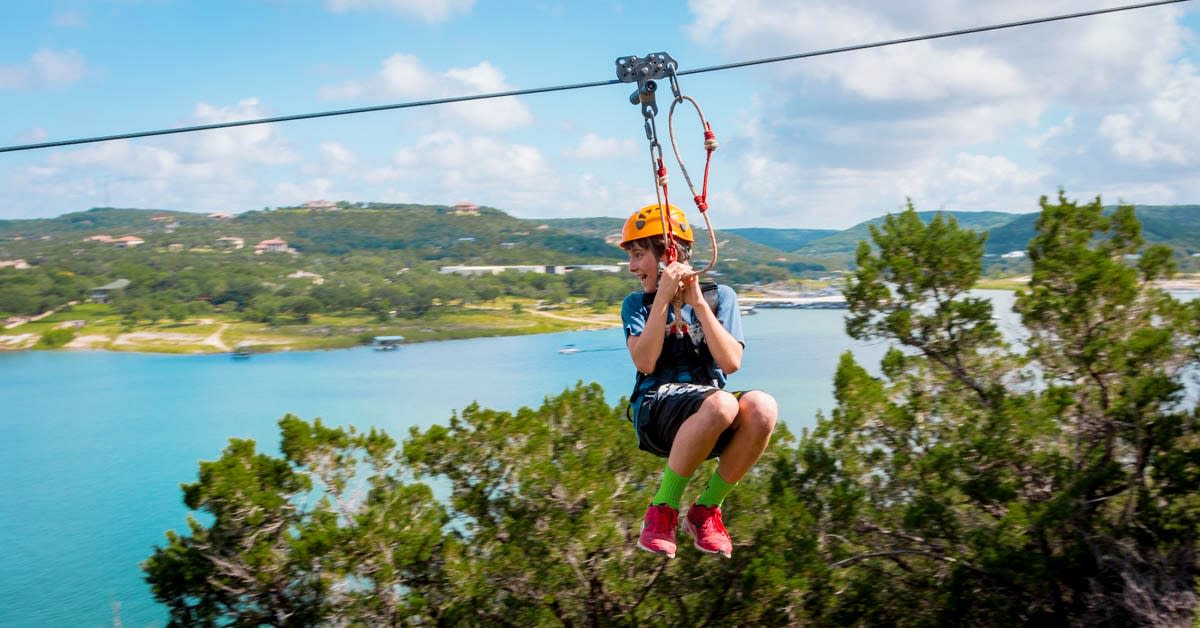 Image of a young boy riding on the zipline with Lake Travis in the background.