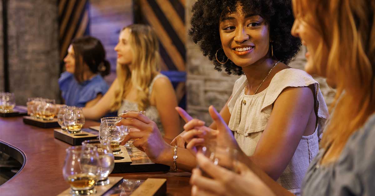 Women at a bar tasting bourbon and talking.