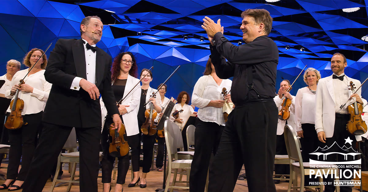 The camera has an on-stage view of an orchestra. All the musicians are dressed in white tops and black pants, holding their instruments. In front of them are two male conductors (?) approaching each other while clapping for their performers.