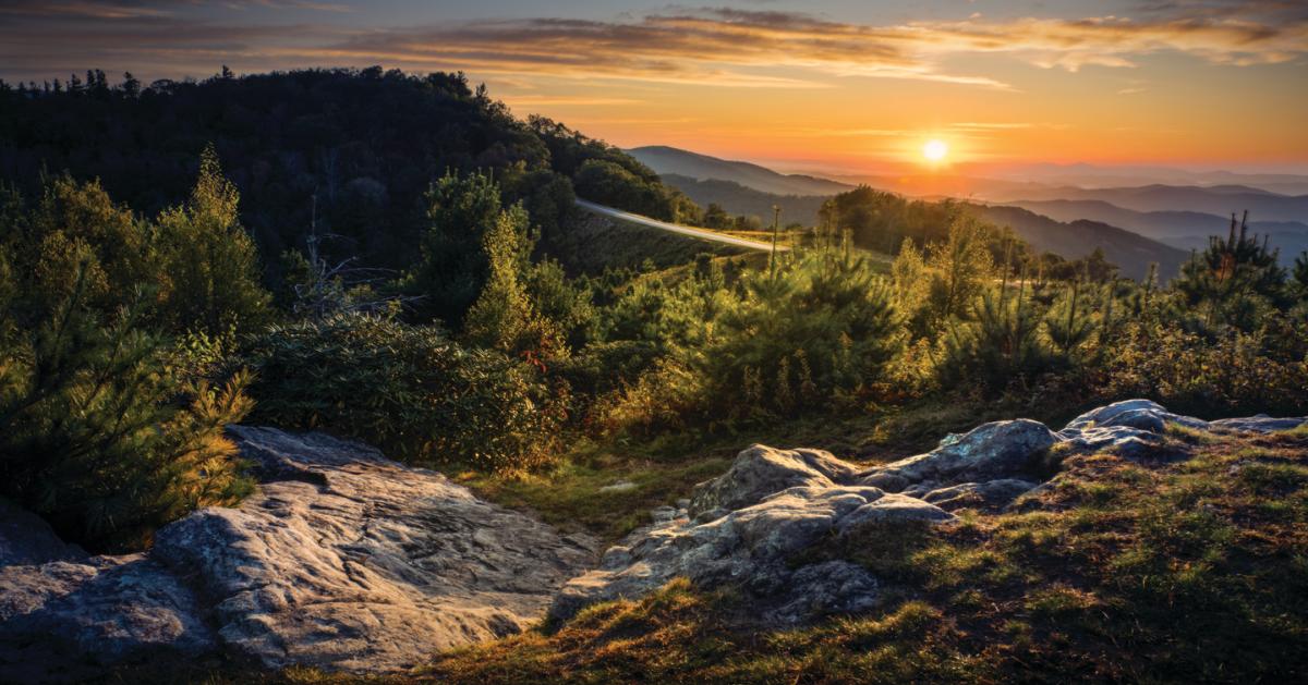 The sun rises along a blue mountain vista, illuminating the Blue Ridge Parkway road in the center of the photograph and a rocky outcropping in the foreground.