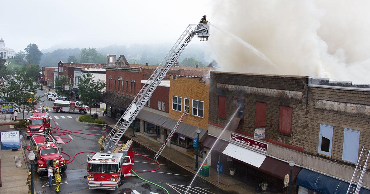 Firefighters on an extended aerial ladder spray water onto a heavily smoking fire on the roof of a brick commercial building. Below, several fire engines are parked on a wet downtown street in front of businesses, including Trevalino's Restaurant. A domed courthouse on a hill is visible in the distant background.