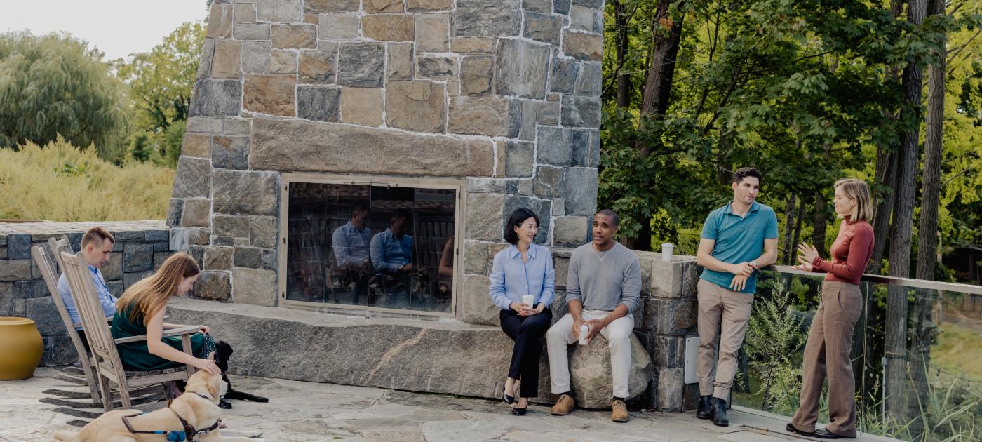 A group of people are outdoors chatting by a large stone fireplace.