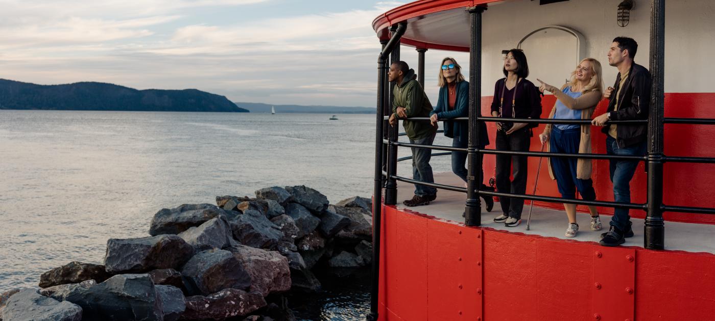 Five people stand on the balcony of the Tarrytown Lighthouse at sunset looking out at the Hudson River.
