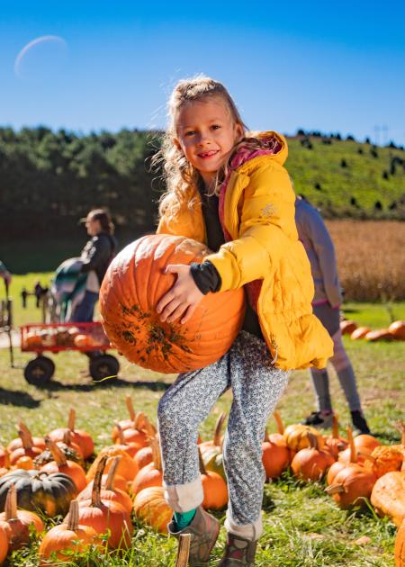 A young girl in a yellow jacket stands in a fields of pumpkins, and holds a large orange pumpkin with both hands while smiling at the camera.