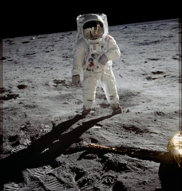 An astronaut in a white spacesuit stands on the Moon during the Apollo 11 mission, with footprints visible in the lunar surface and the blackness of space stretching across the horizon.