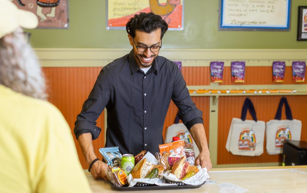 Man in navy blue button up shirt smiles as he picks up and looks at a tray holding sandwiches, bagged chips, and food items.
