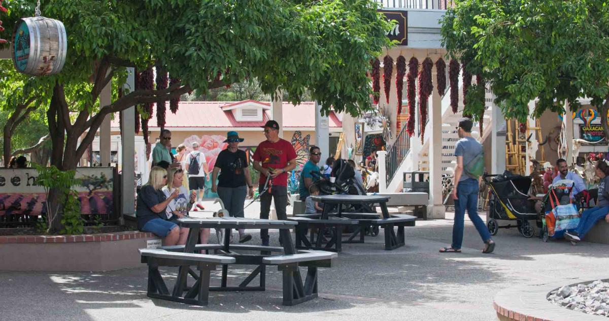 People relaxing and walking around an outdoor market area with picnic tables, trees, and shopfronts in the background.