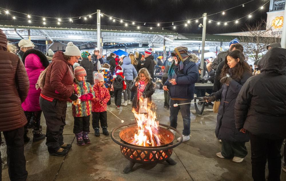 people of all ages stand outside on concrete circled around an active firepit, some children toast marshmellows on a stick over the fire as one person assists.  Others stare at the fire