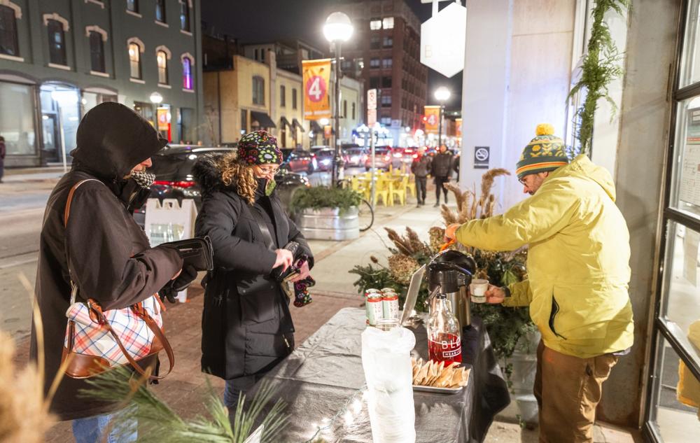 Man in coat and hat stands outside building and behind table, pouring samples of a beverage for two people wearing coats standing in line.