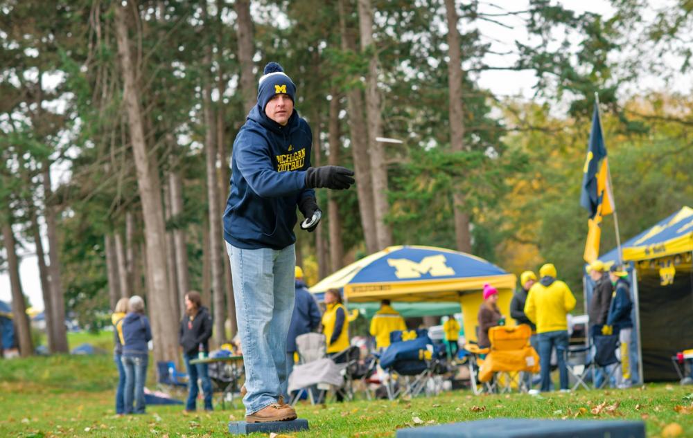 Man wearing navy blue and yellow sweatshirt and snow hat holds an item mid-throw, part of yard games during a tailgate.