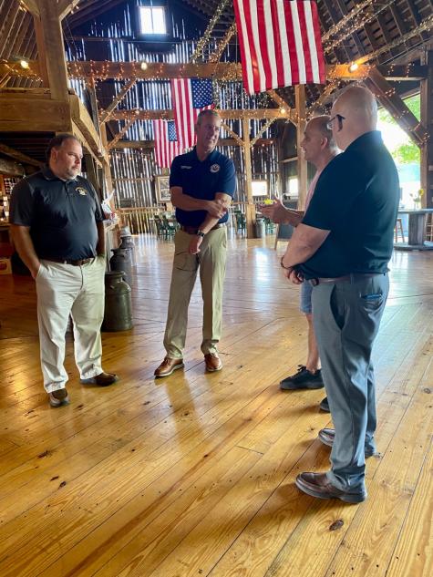 NYS Assemblymen meeting with Muranda Cheese owner inside of a rustic wooden barn surrounded by string lights and American flags.