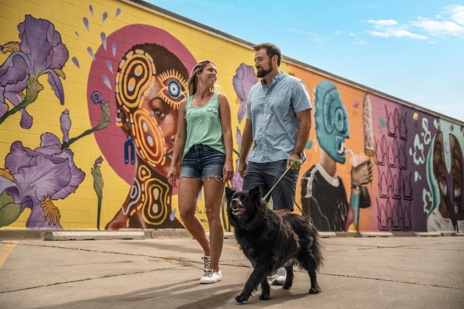 A couple walks in front of an art mural with their dog downtown