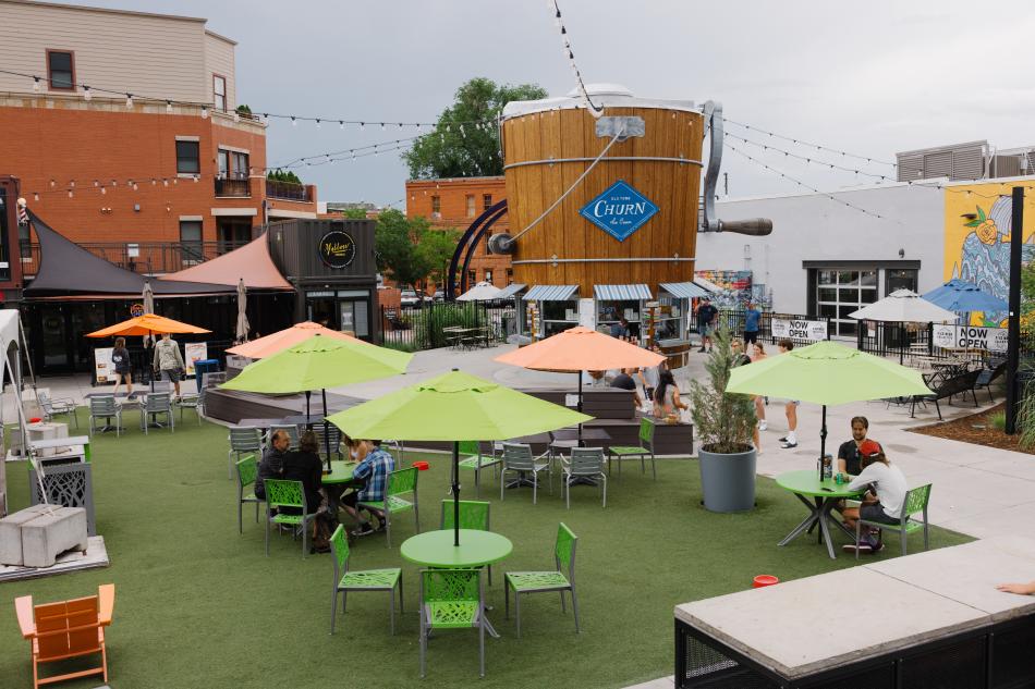 A central gathering place with picnic tables and with a building shaped like an ice cream churn in the middle with twinkle lights going off to other buildings.