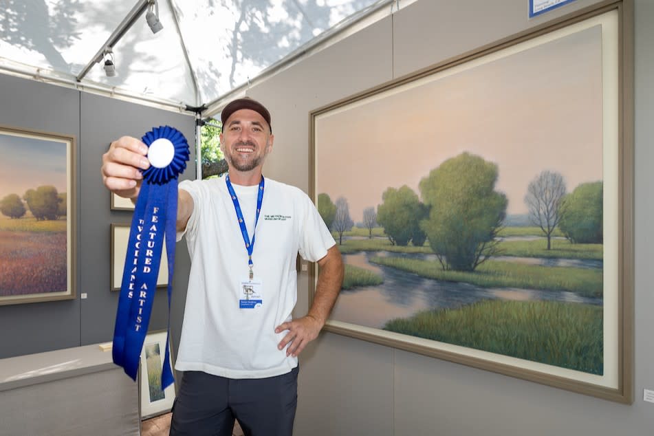 A man poses with his watercolor (?) paintings at the 2025 Waterway Arts Festival, holding a bright blue "Featured Artist" ribbon to the camera. The man is wearing a white T-shirt, gray shorts, a brown ballcap, and a blue WWAF lanyard. The paintings are of a marshy landscape at sunset. Both the colors and textures are subtle and soft.