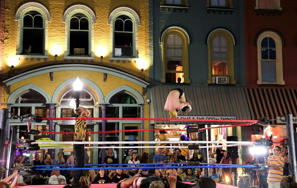 Wrestling ring sits outside with colorful buildings in the background. Within the wrestling ring, a wrestler does a cannonball and is mid-air. The referee watches with his hands on his head.