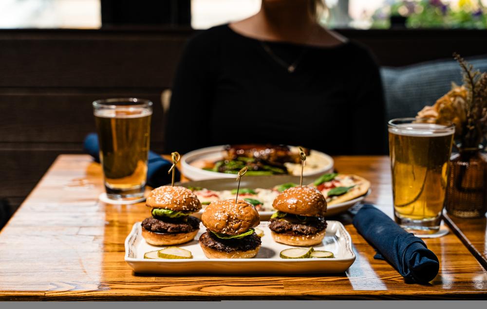 The viewer and a young woman are seated across each other at Bosscat Kitchen. On the table are two glasses of beer, a plate of sliders, and a chicken and vegetable dish.