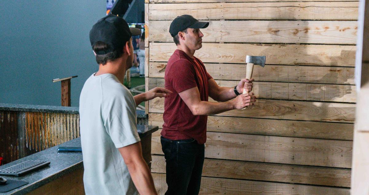 Two men participate in an axe-throwing activity, with one man preparing to throw while the other watches.