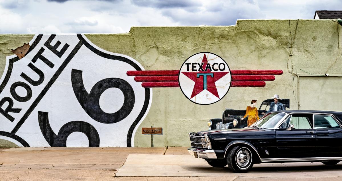 A vintage car parked beside a wall with Route 66 and Texaco logos. A person in retro clothing stands near an old car.