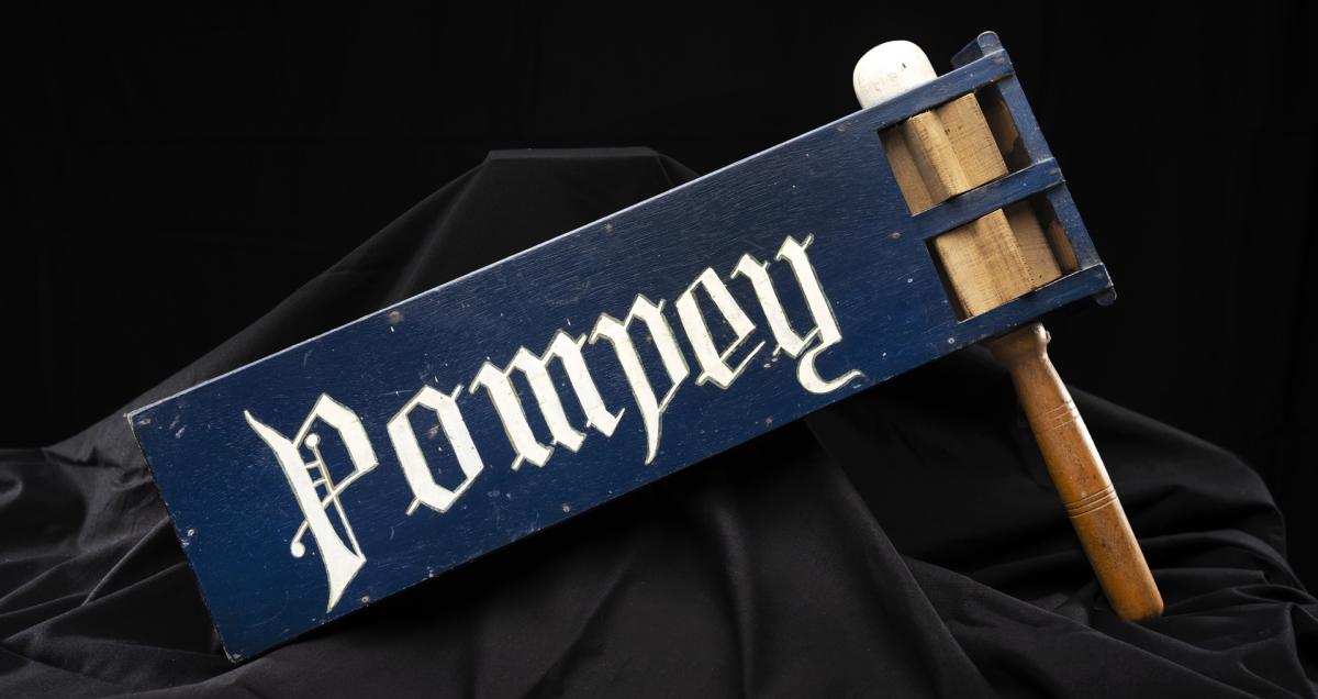 A wooden rattle used at a football match. It has a wooden handle and a blue section with the word Pompey in white font.