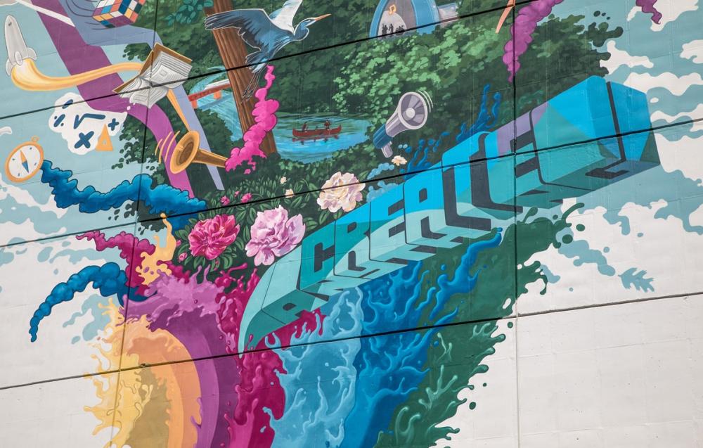 section of a colorful mural that spells the word "create" and underneath transforms to the word "anything". Behind the words are a colorful explosion of waves that transform into activites like a trumpet, flowers, math problems. Items that represent Ann Arbor