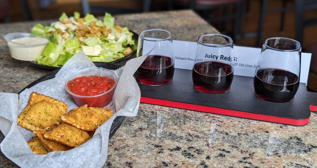 The image is of a red wine flight featuring 3 small glasses of red wine sitting next to a small Ceasar salad and cheese ravioli.