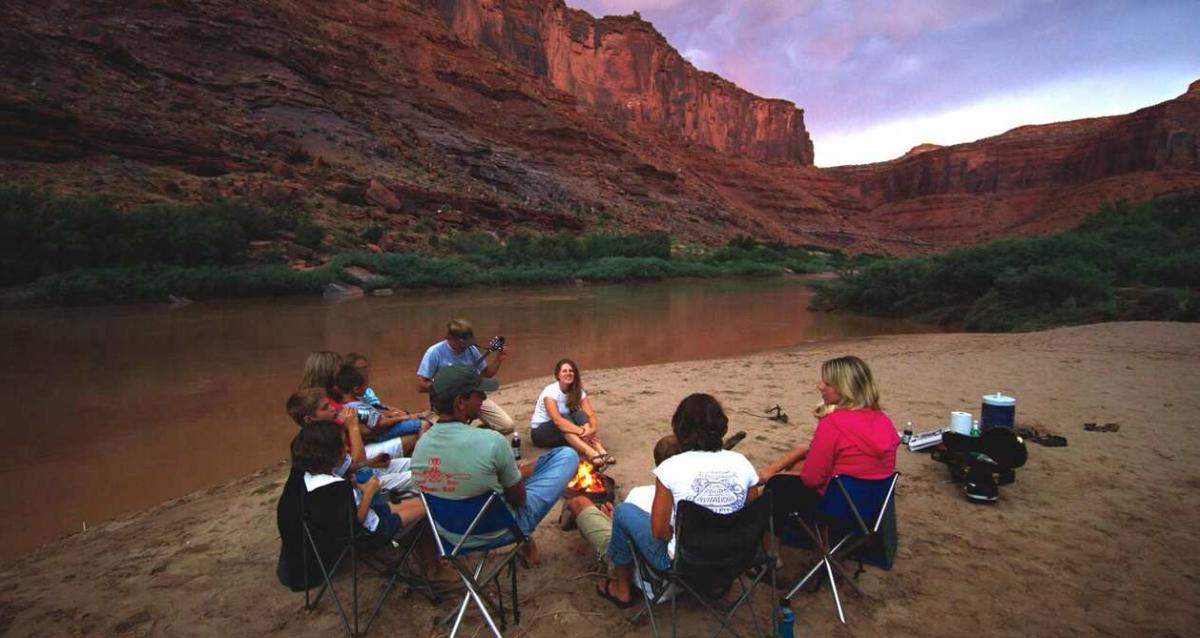 Family sitting on camp chairs around a campfire by the river with a guy playing guitar