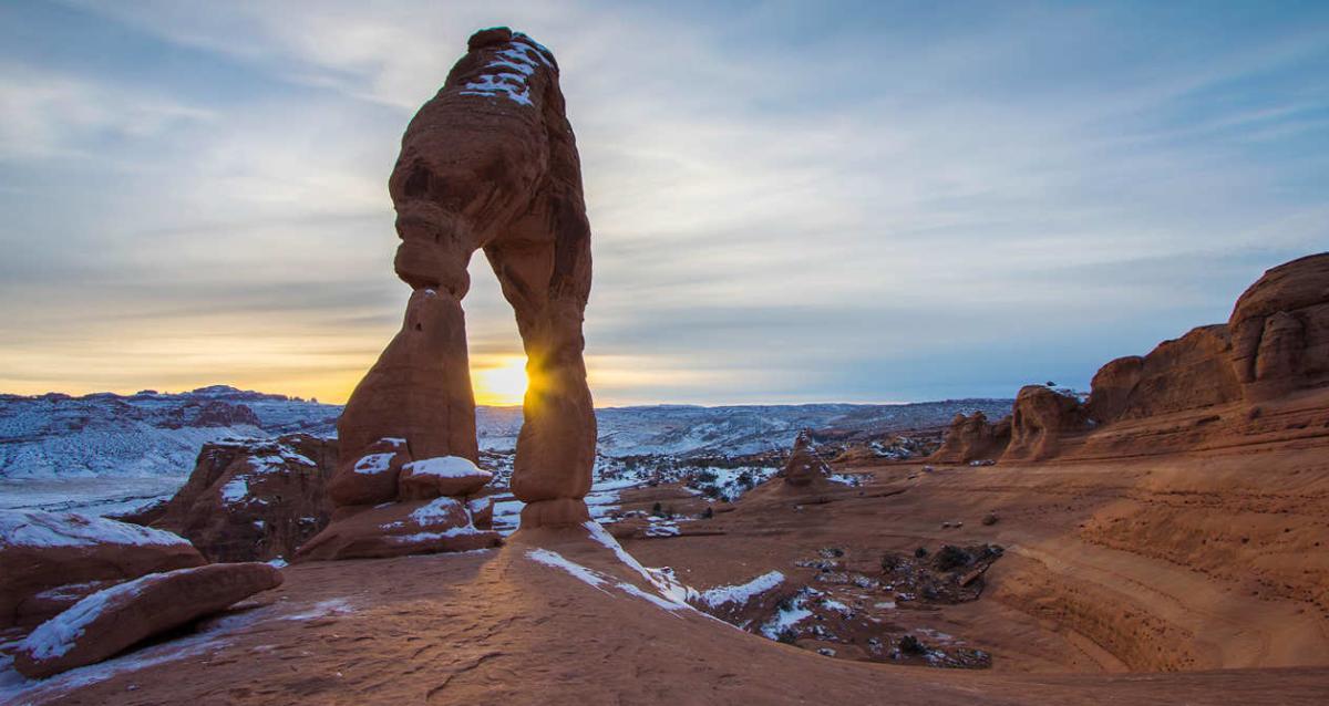 View of the sunset through Delicate Arch with snow on the ground.