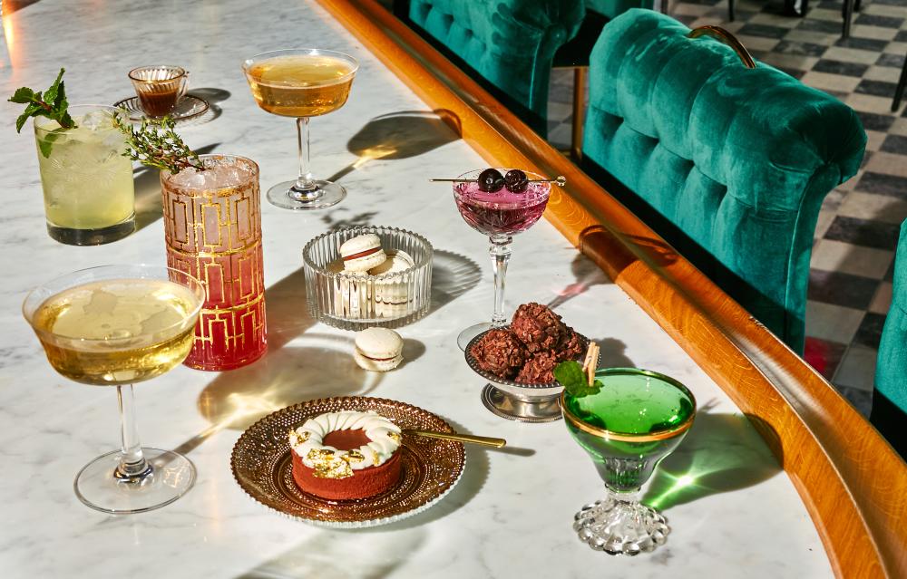 Vintage-style drink glasses surrounding desserts on gilded serveware on a marble bar next to velvet barstools.