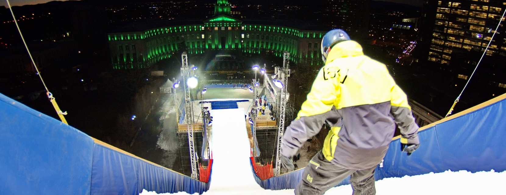 A snowboarder is set at the top of a massive slope with the Denver capitol facing them