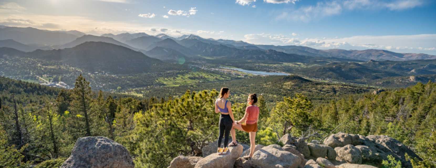 Two people gaze out at a mountain vista surrounded by green trees in Estes Park