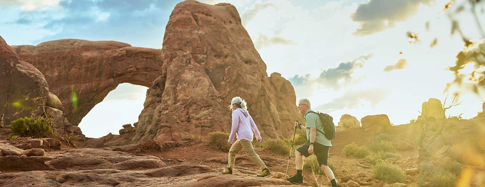 Two people are hiking by an arch at Arches National Park near Moab, Utah.
