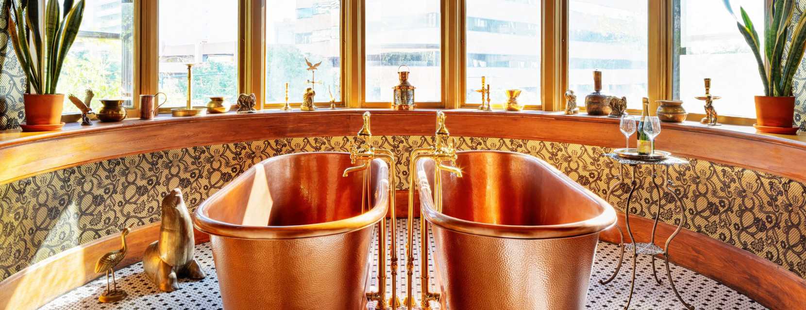 Two bathtubs sitting next to each in front of windows along with a table holding a wine bottle and two glasses at the gilded-age hotel, Urban Cowboy Denver.
