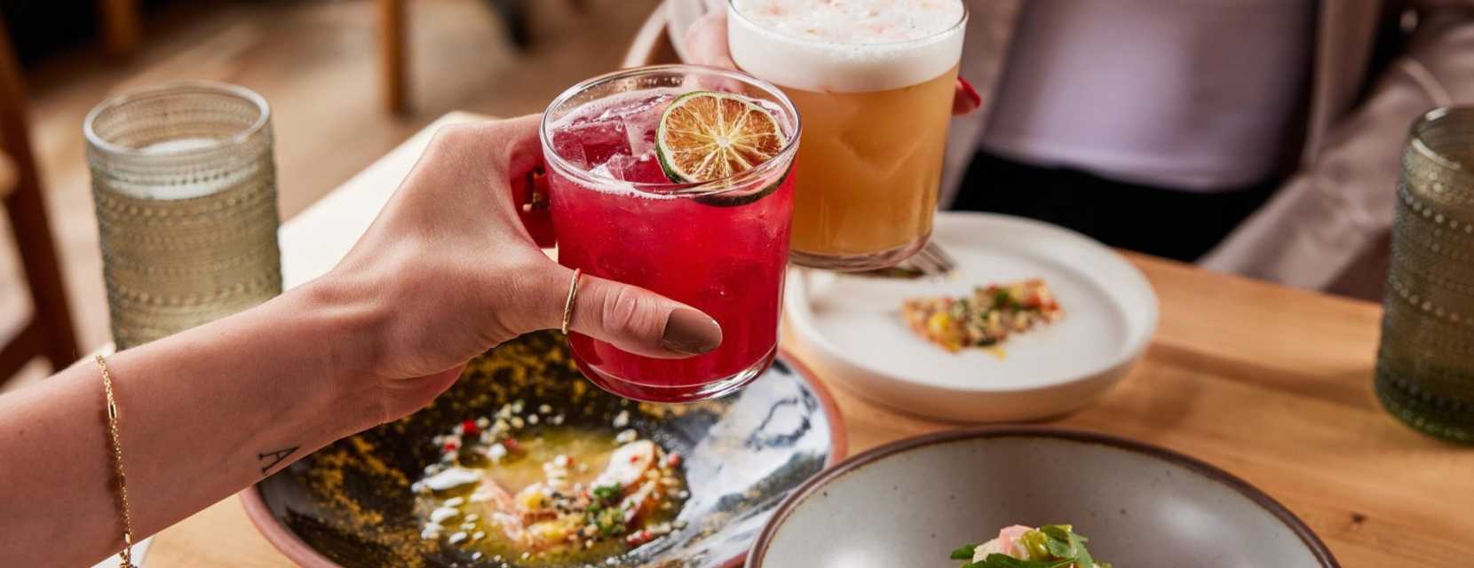 Two people cheers colorful cocktails with plates below at Alma Fonda Fina in Denver