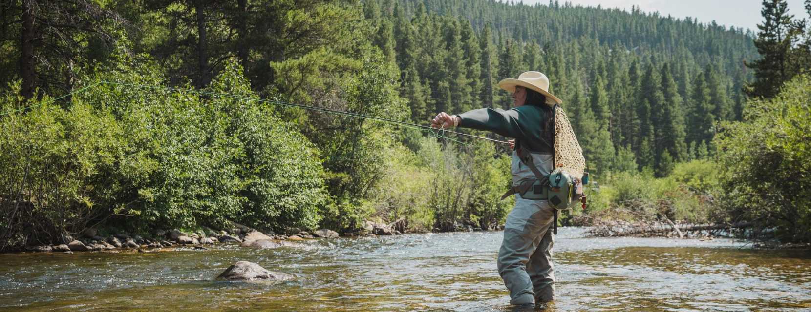 An angler stands in a river near Denver fly-fishing with pine trees in the background