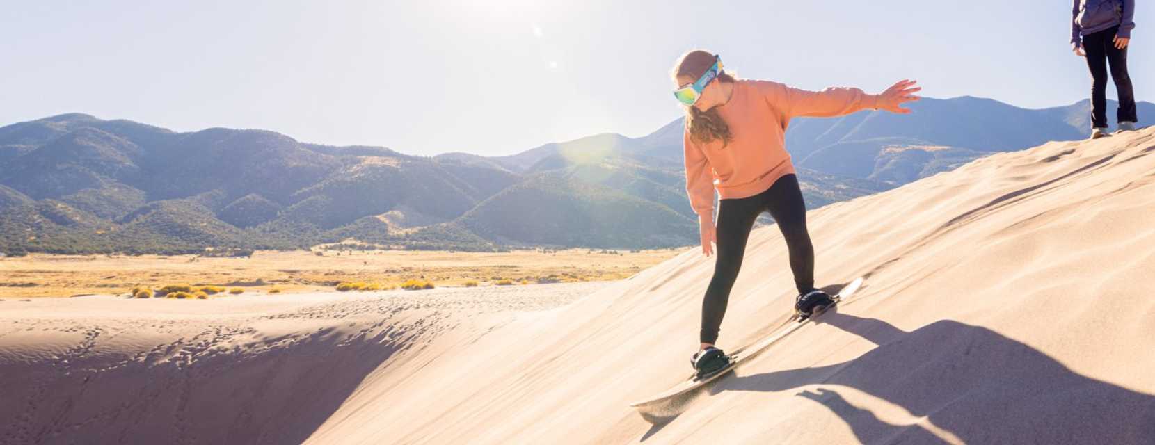 A child wearing goggles sandboards down a dune at Great Sand Dunes National Park & Preserve with mountains in the backgroun