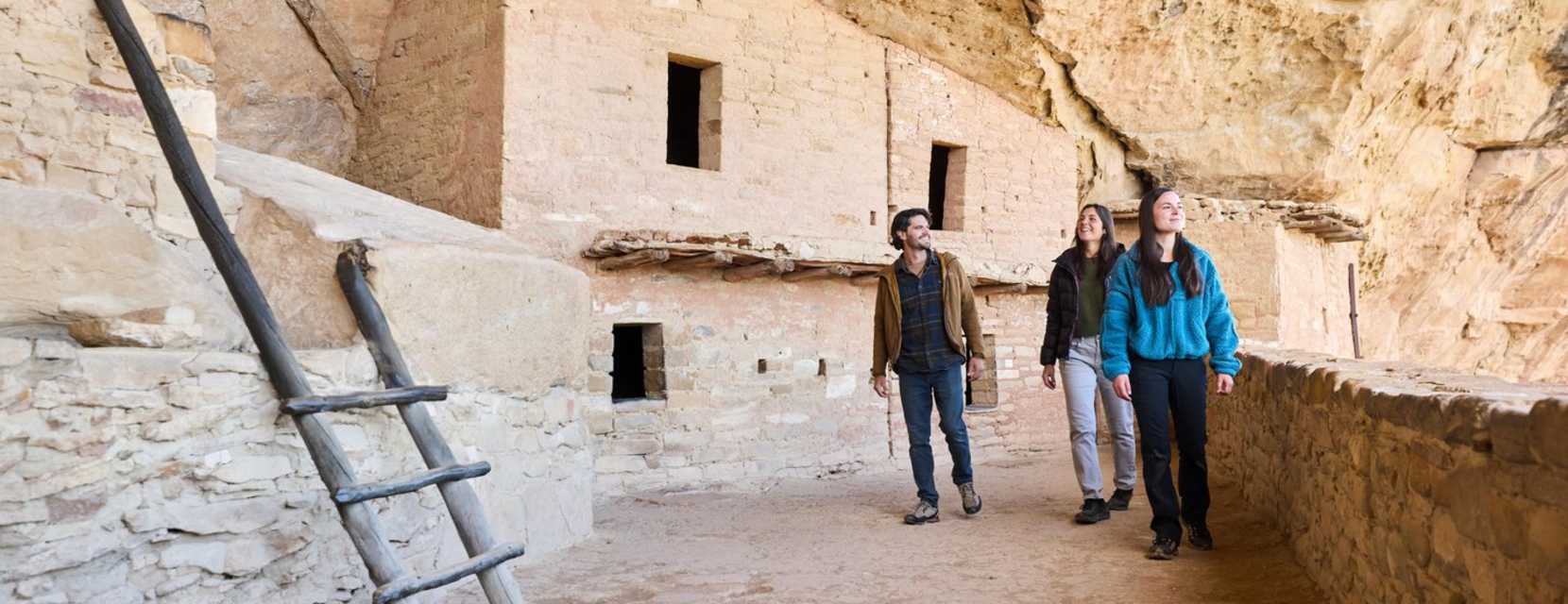 Three people gaze around at a cliff dwelling at Mesa Verde National Park. Tan stone walls are dotted with windows