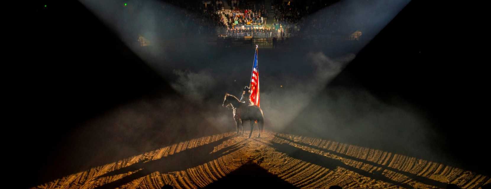 A horseback rider holding a flag is in the center of the arena with spotlights facing them