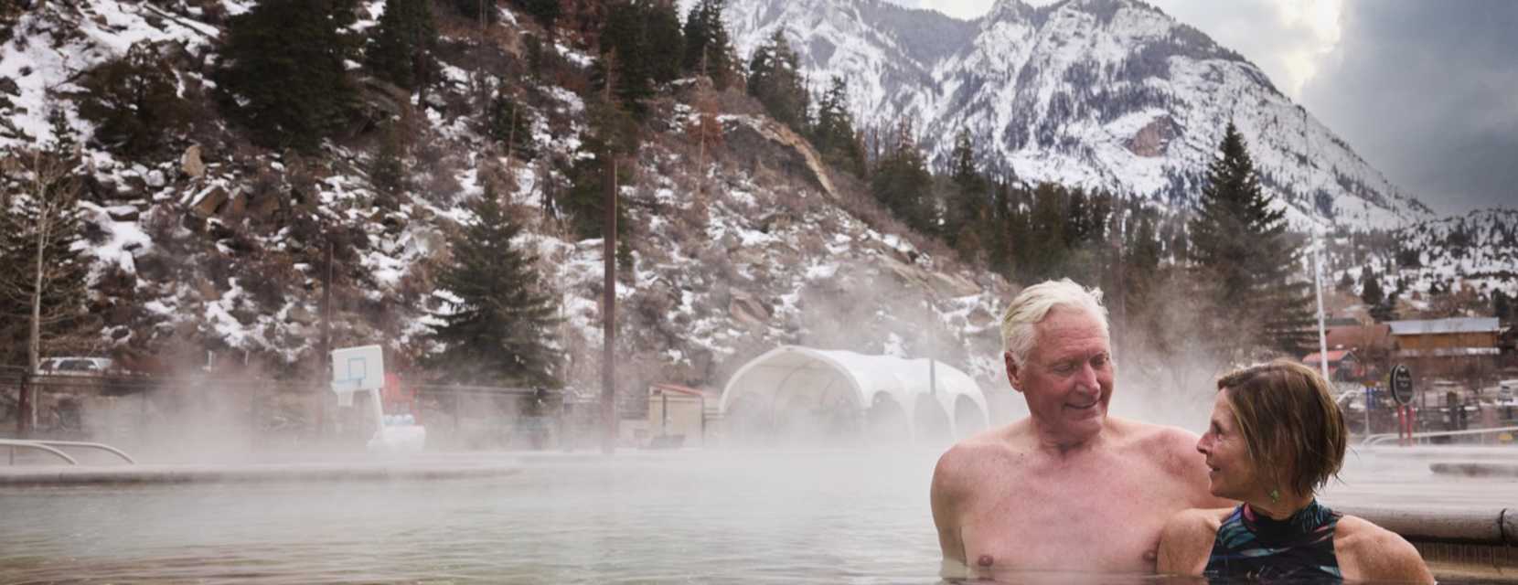 Two people smile at eachother while sitting in a hot springs pool with a snow-covered mountain in the background