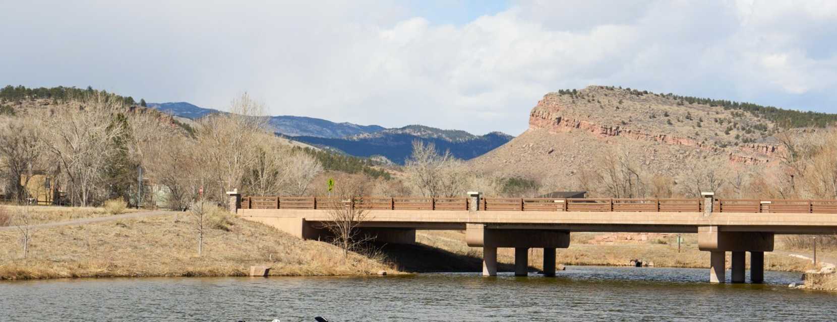 Two people kayak along St. Vrain Creek wiht mountains and a bridge in the background