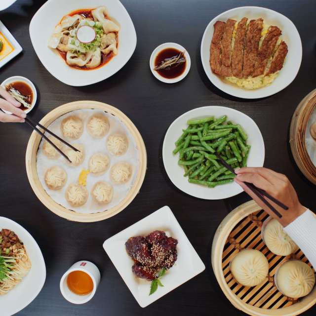 Black wood table full of brightly colored food, and dumplings in wooden baskets, hands reaching across the table with chopsticks