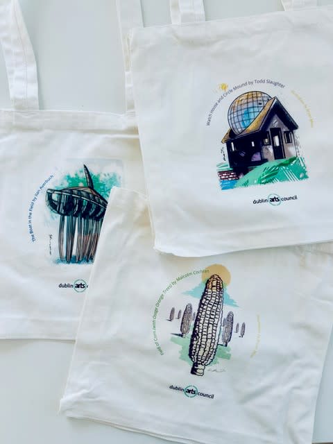 Three white canvas tote bags featuring artistic illustrations of public art installations in Dublin, Ohio, designed for the Dublin Arts Council's 2025 Art Pass. Each bag showcases a different artwork, including a corn sculpture, a spherical glass house, and a modern metal structure, with accompanying text describing the installations.