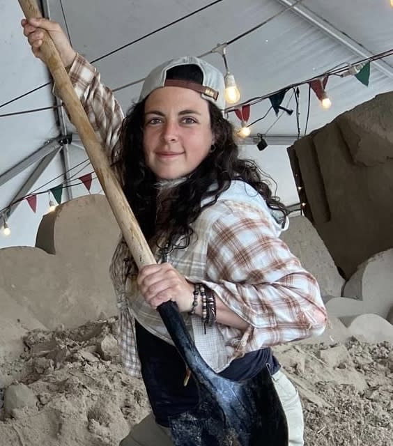 A woman wearing a baseball cap and a flannel shirt holds up a shovel like a jousting weapon, smiling at the camera in front of a half-carved pile of sand.